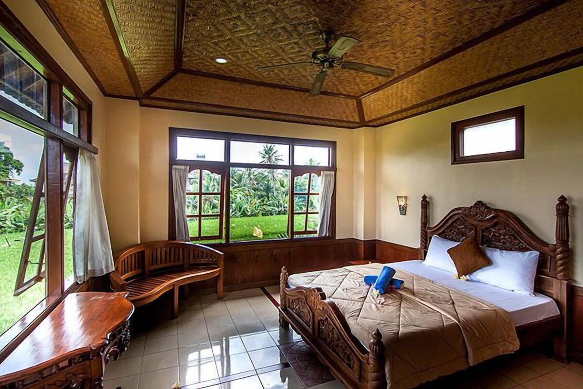 A bedroom with a wooden bed, a wooden bench, a ceiling fan, and large windows with a view of greenery.
