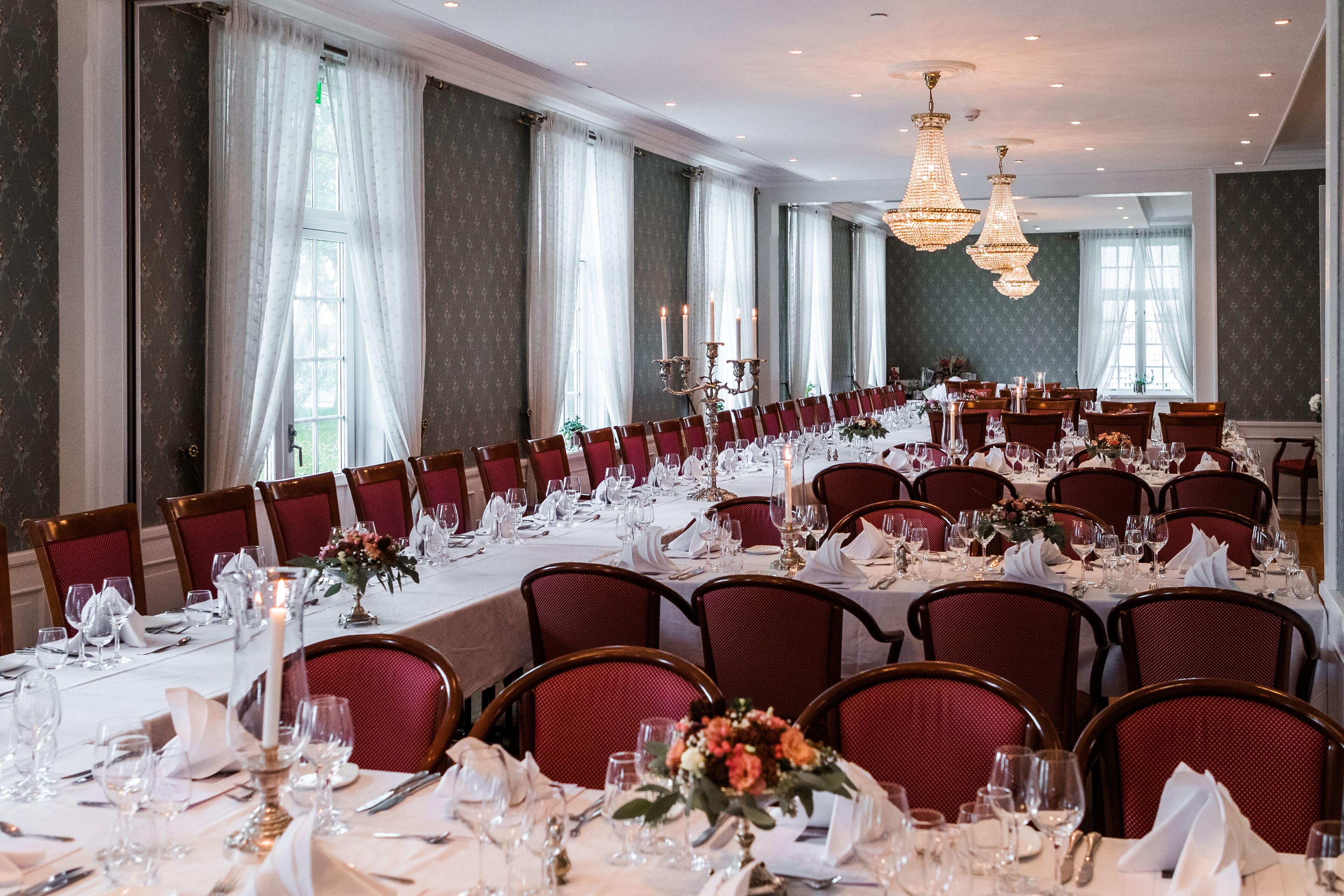 A banquet hall with long tables set for a formal event, featuring chandeliers and red upholstered chairs.