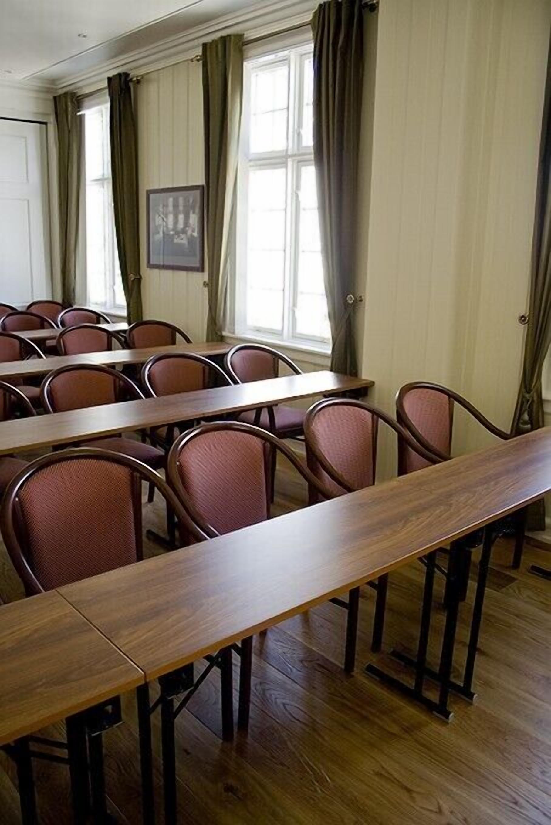 Une salle de conférence avec de longues tables et des chaises, de grandes fenêtres avec des rideaux et un tableau encadré au mur.