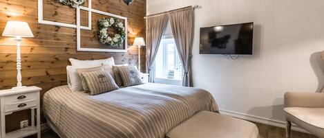 A bedroom with a wooden paneled wall, a bed with striped bedding, a bedside table with a lamp, a TV mounted on the wall, and a window with curtains.