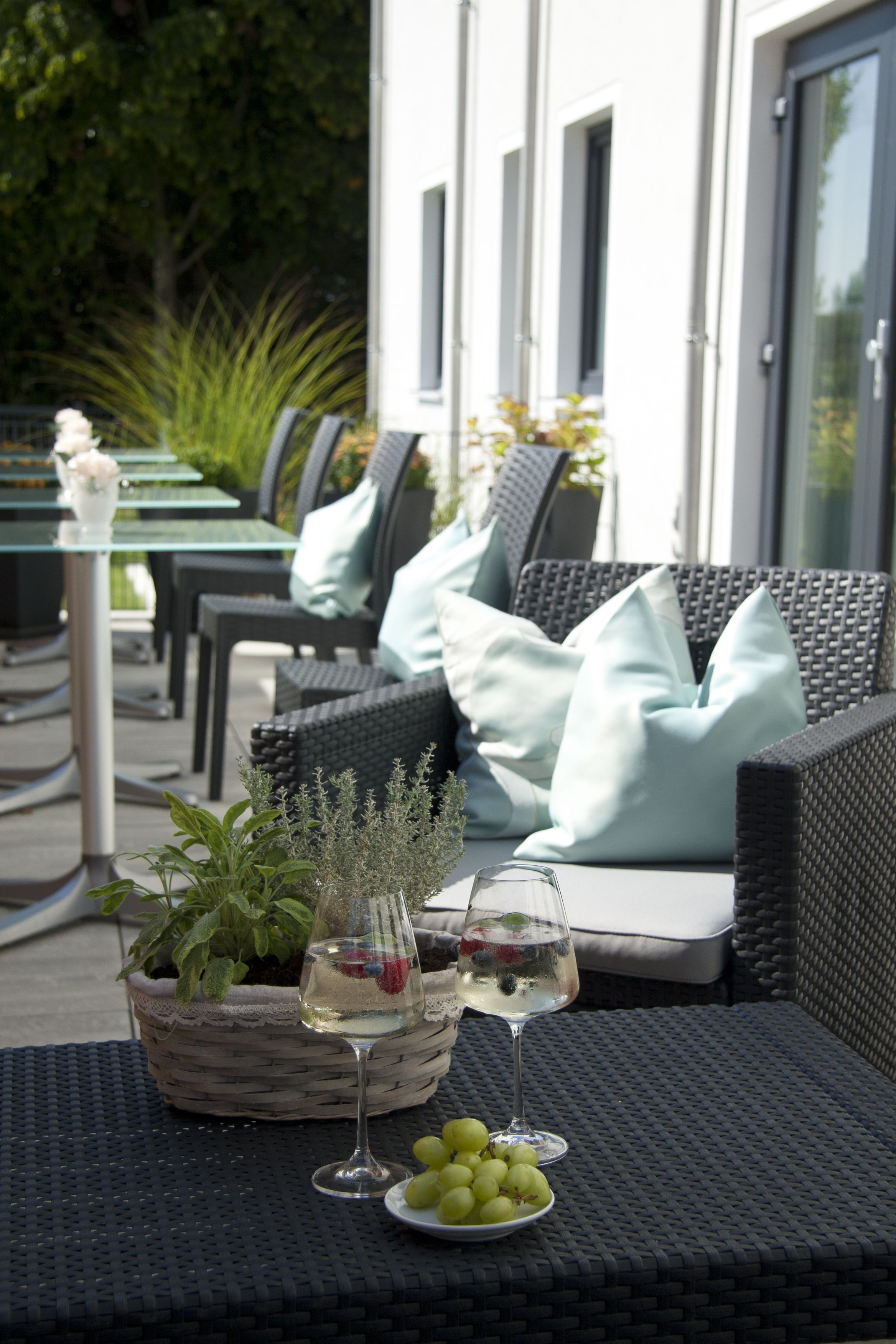 Een terras met rieten meubels, een tafel met een potplant en een schaal druiven, en twee wijnglazen.