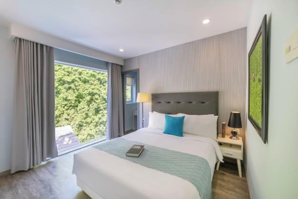 Quarto de hotel com uma cama grande, uma mesa de cabeceira com abajur, uma janela com cortinas e um quadro na parede.