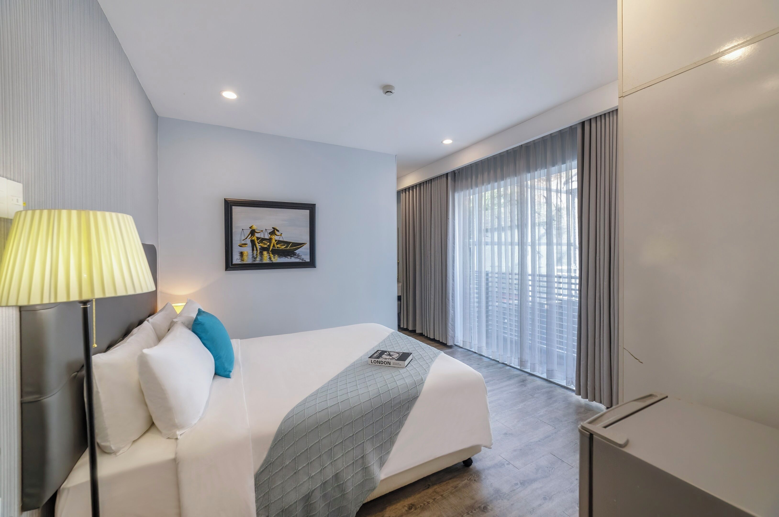 Habitación de hotel con una cama, una lámpara, un cuadro enmarco y un ventanal con cortinas.