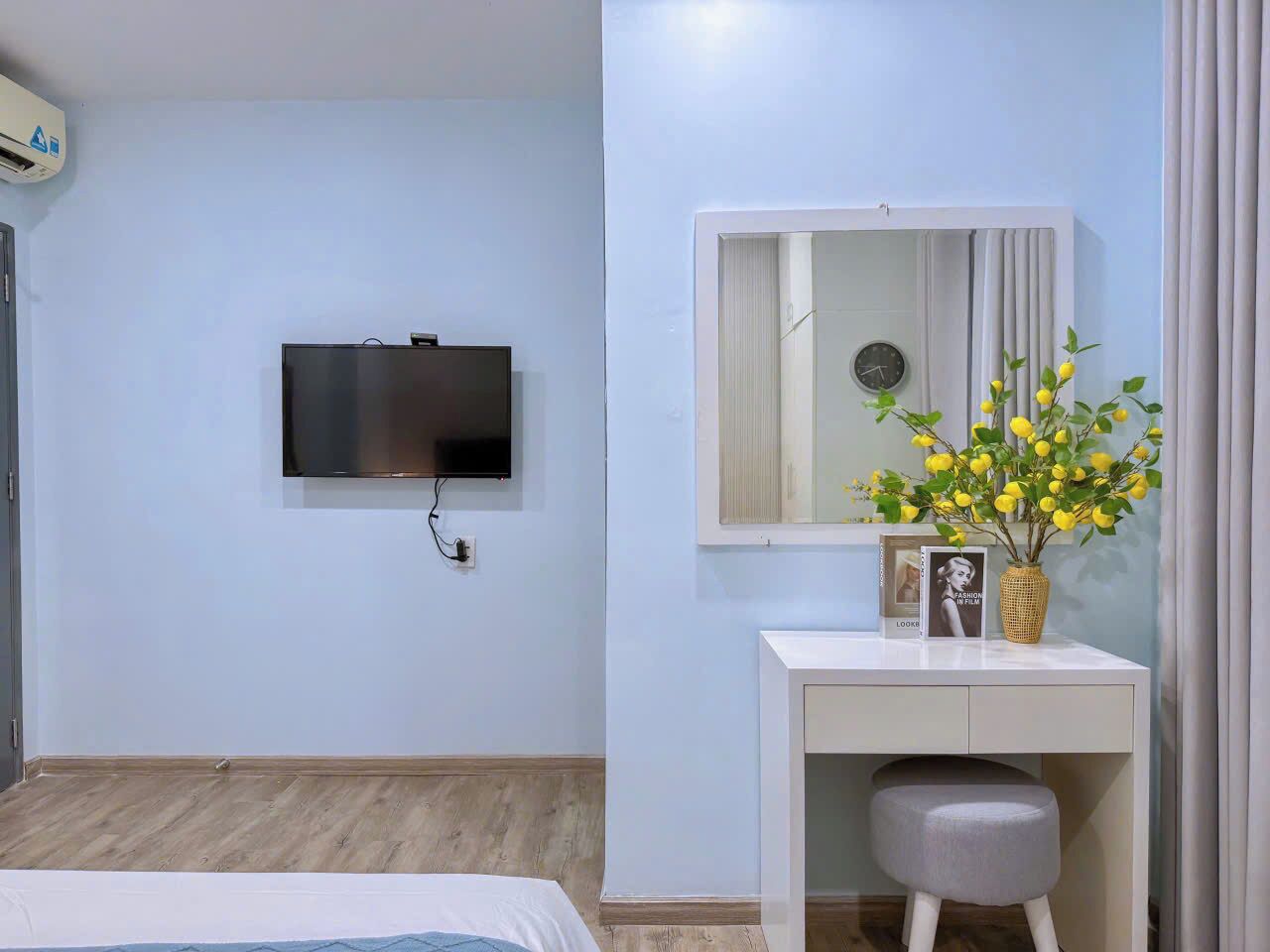 A hotel room with a flat-screen TV mounted on the wall, a small desk with a mirror, a vase with yellow flowers, and a bedside table.