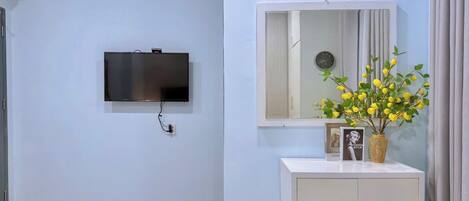 Habitación de hotel con una televisión de pantalla plana montada en la pared, un pequeño escritorio con un espejo, un jarrón con flores amarillas y una mesita de noche.