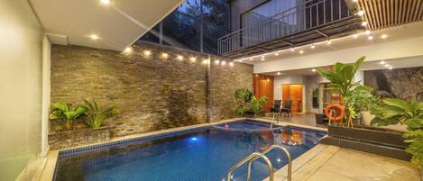 An indoor swimming pool with a stone wall, a balcony, and a seating area with plants.