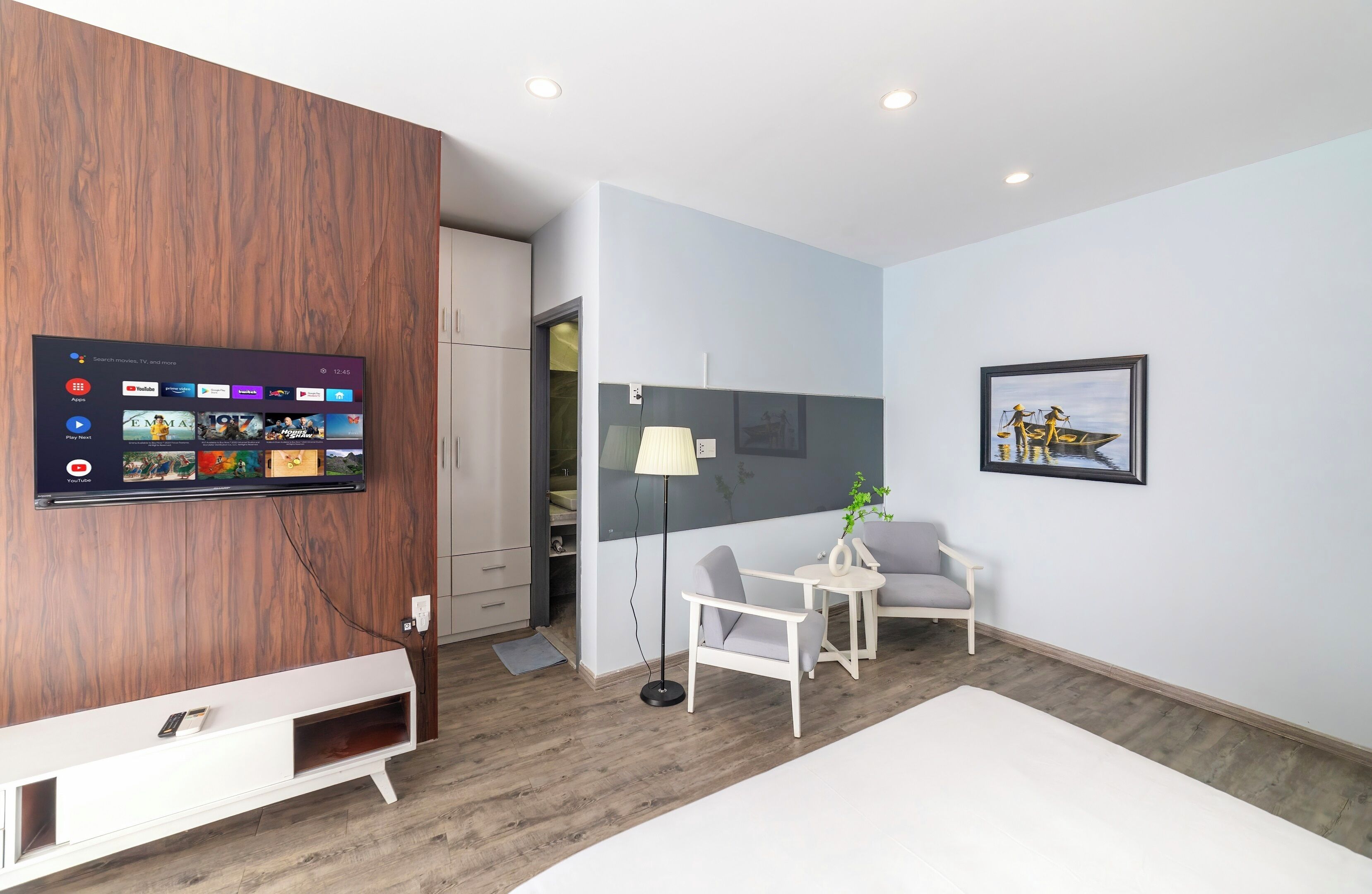 Una habitación moderna con un televisor de pared mostrando la interfaz de un servicio de streaming, un tocador blanco, una pequeña mesa blanca con una silla y una obra de arte enmarcada en la pared.