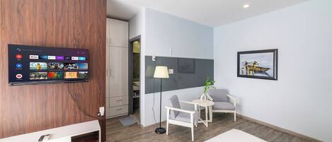 Una habitación moderna con un televisor de pared mostrando la interfaz de un servicio de streaming, un tocador blanco, una pequeña mesa blanca con una silla y una obra de arte enmarcada en la pared.