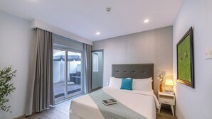 A hotel room with a large bed, a nightstand with a lamp, a framed picture on the wall, and a sliding glass door leading to an outdoor area. - Hese Boutique Hotel & Apartment (Ho Chi Minh City)