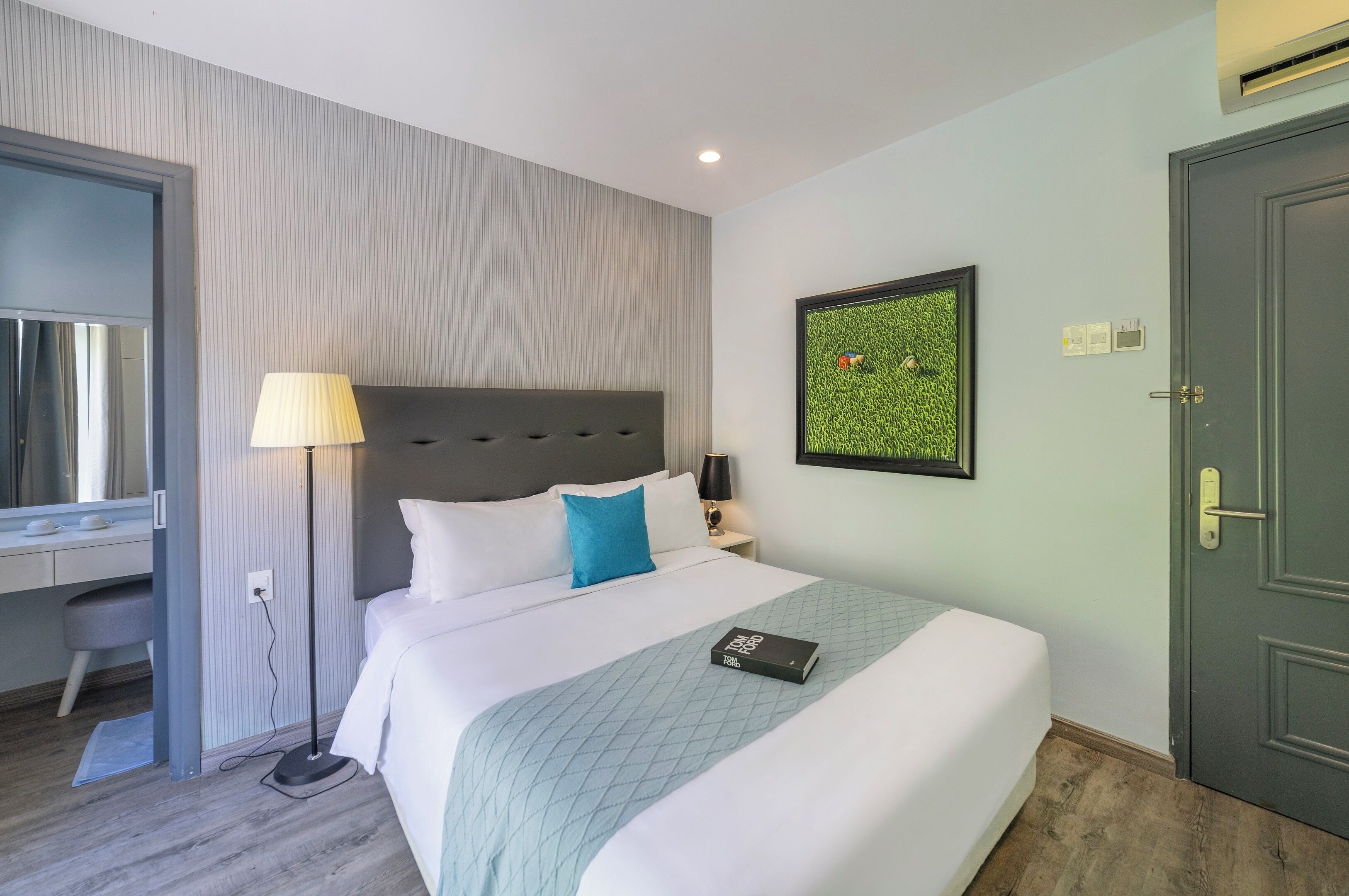 Una cama bien hecha con un cojín azul, una lámpara de noche y un cuadro en la pared.