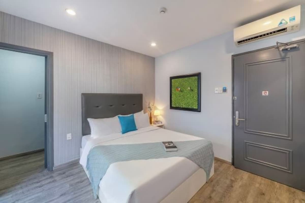 A hotel room with a bed, a nightstand, an air conditioning unit, and a door.
