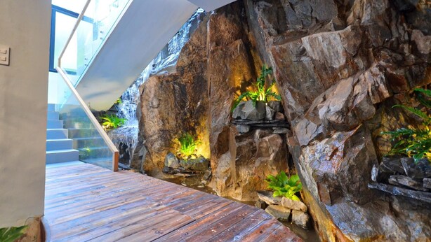 A modern interior with a wooden deck, a stone wall with a waterfall feature, and a staircase leading upwards. - Hese Boutique Hotel & Apartment (Ho Chi Minh City)