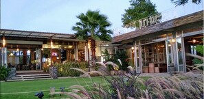 A modern building with a covered patio area, outdoor seating, and a well-maintained garden with palm trees and ornamental grasses.