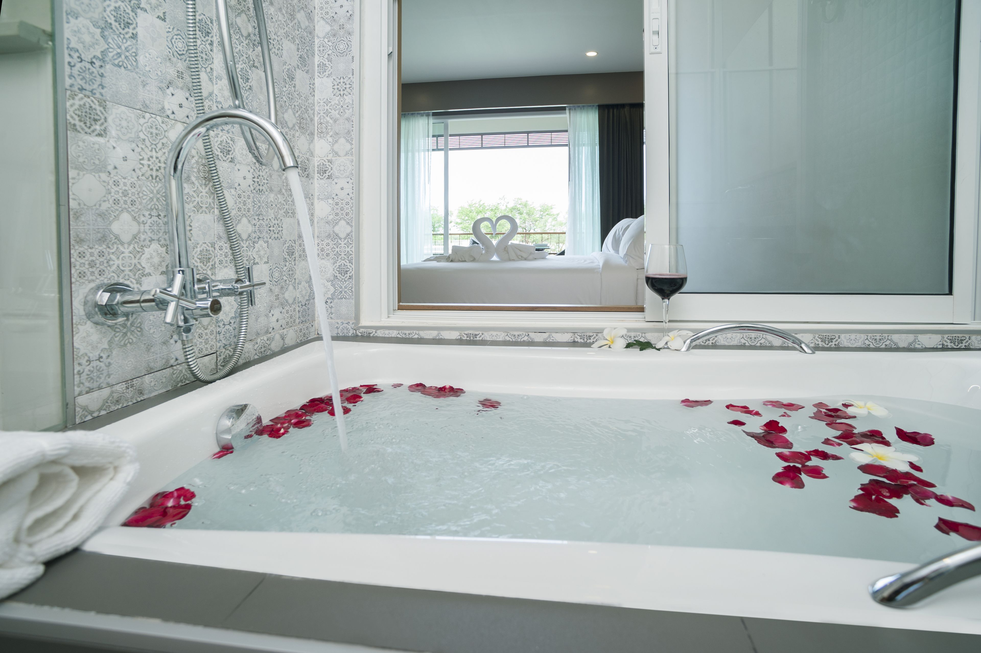 A bathtub with rose petals and a glass of wine, a showerhead, and a view of a balcony with a heart-shaped decoration.