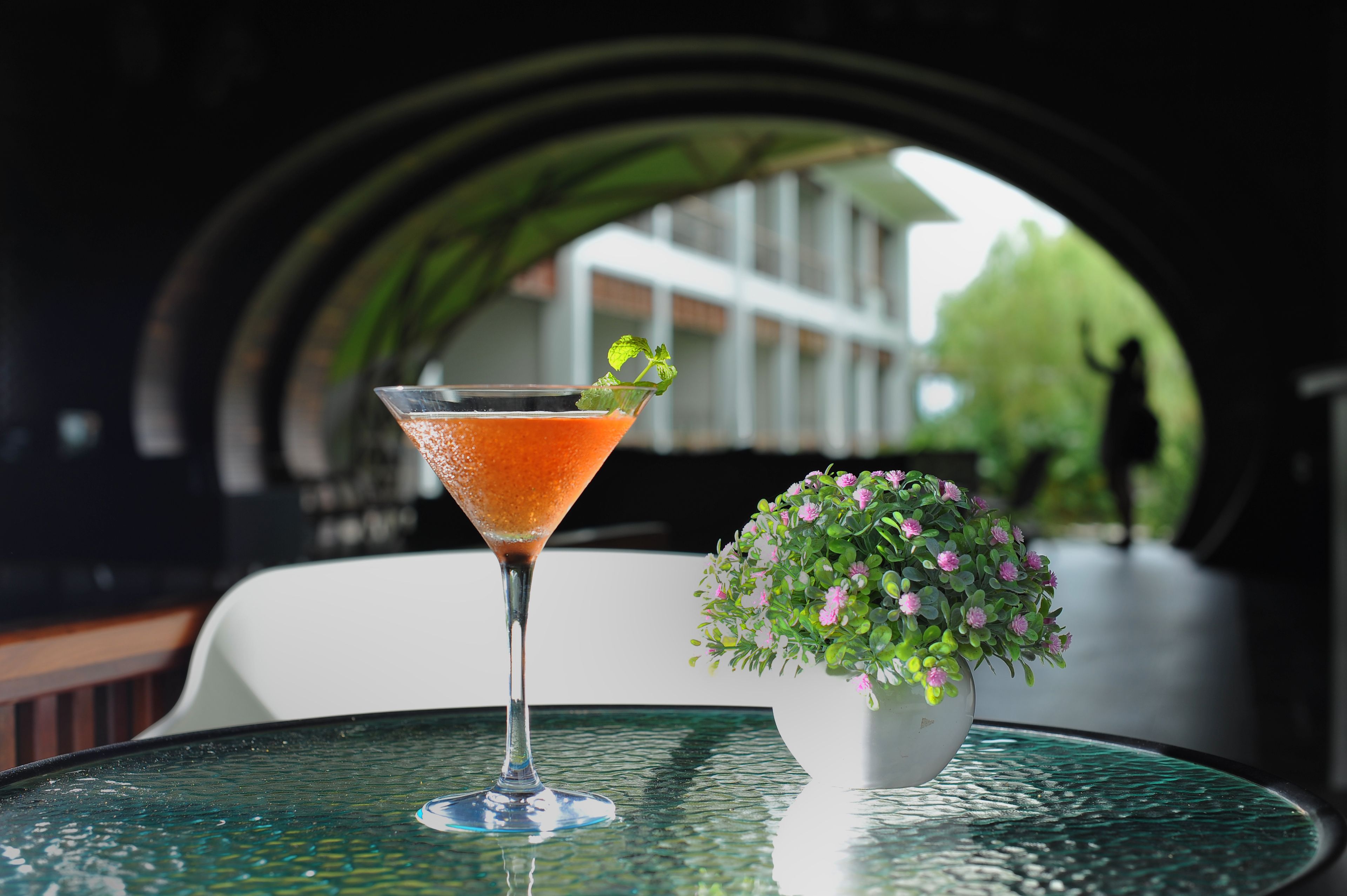 A martini glass with a citrus garnish on a glass table, a small potted plant with pink flowers, and a view of a building through an archway.