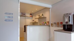 Cozinha moderna com geladeira, máquina de café e uma prateleira com diversos itens. Há placas indicando comodidades como academia, bebidas e lanches.