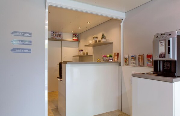 Eine moderne Küche mit einem Kühlschrank, einer Kaffeemaschine und einem Regal mit verschiedenen Gegenständen. Schilder weisen auf Annehmlichkeiten wie ein Fitnessraum, Getränke und Snacks hin.