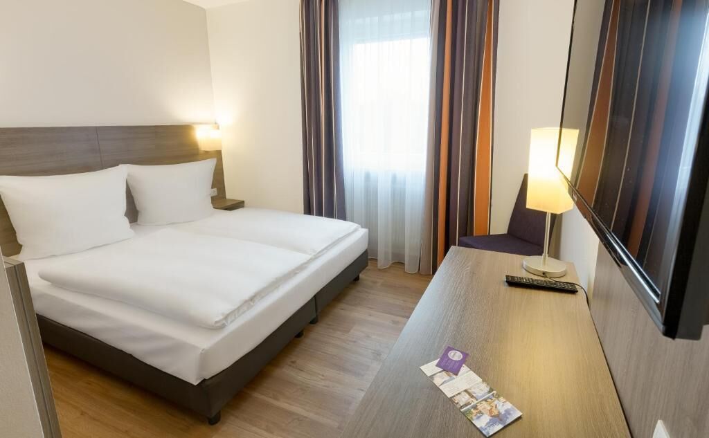 Ein Hotelzimmer mit Bett, Schreibtisch mit Lampe, Fernseher und einem Fenster mit Vorhängen.