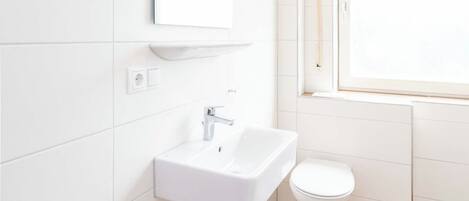Un baño moderno con azulejos blancos, un lavamanos blanco, un inodoro y una ventana.