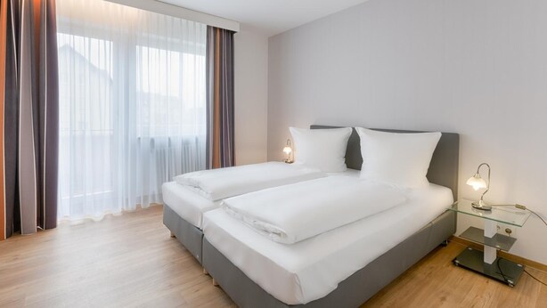 Quarto de hotel com duas camas, uma mesa lateral de vidro e uma luminária de cabeceira.