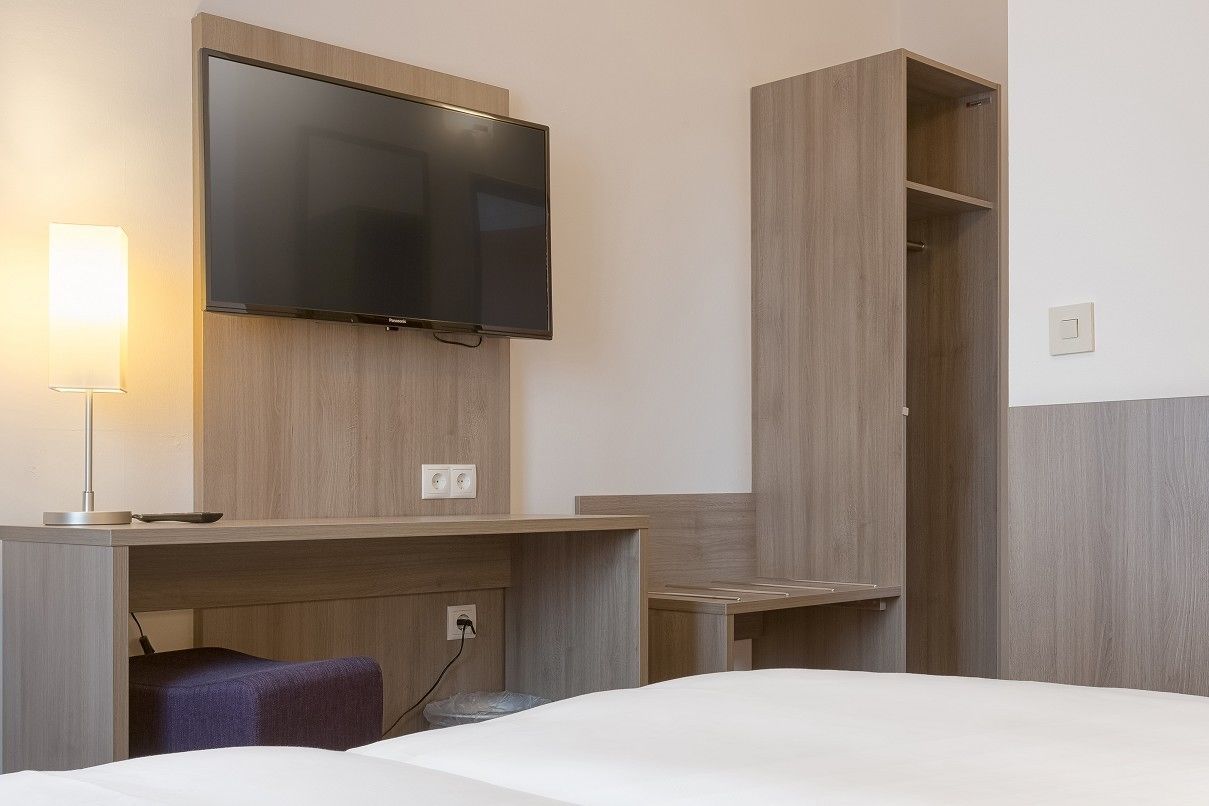 A hotel room with a bed, a desk with a flat-screen TV, a lamp, and a wardrobe.