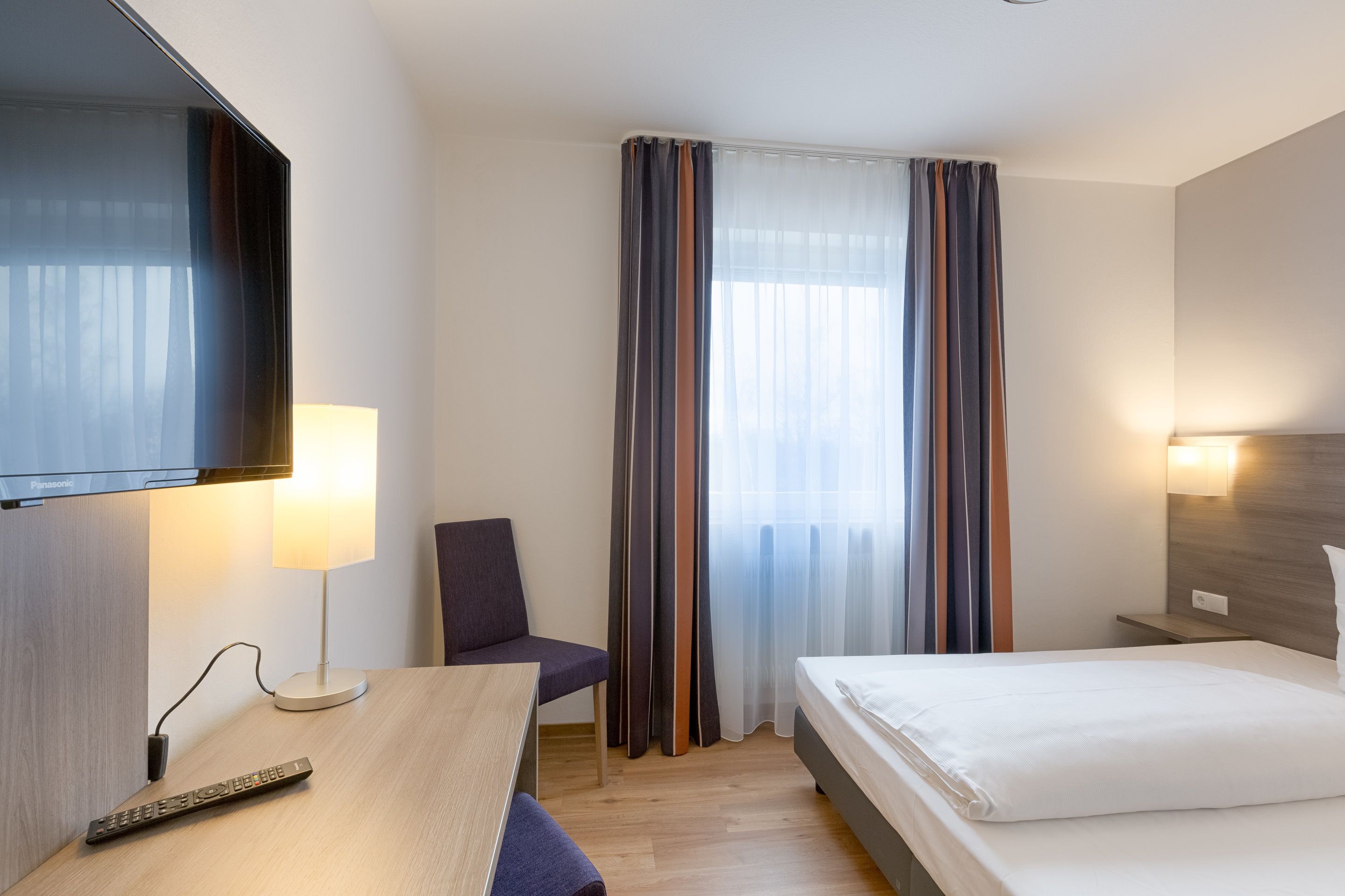 A hotel room with a bed, a desk with a lamp, a chair, a TV mounted on the wall, and a window with curtains.