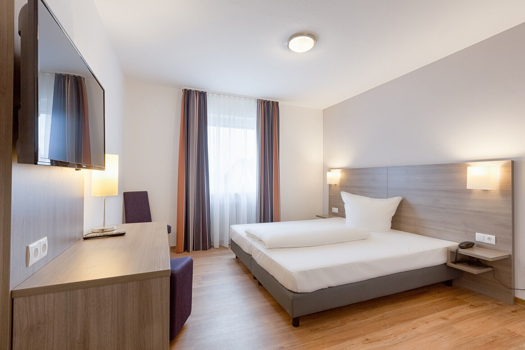 Ein modernes Hotelzimmer mit einem großen Bett, einem Schreibtisch mit Stuhl, einem Flachbildfernseher und einem Nachttisch mit Lampe.