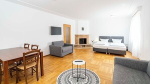 Um quarto com uma mesa de jantar de madeira, dois sofás cinzentos, uma cama com dois colchões brancos, uma lareira e uma televisão presa à parede.