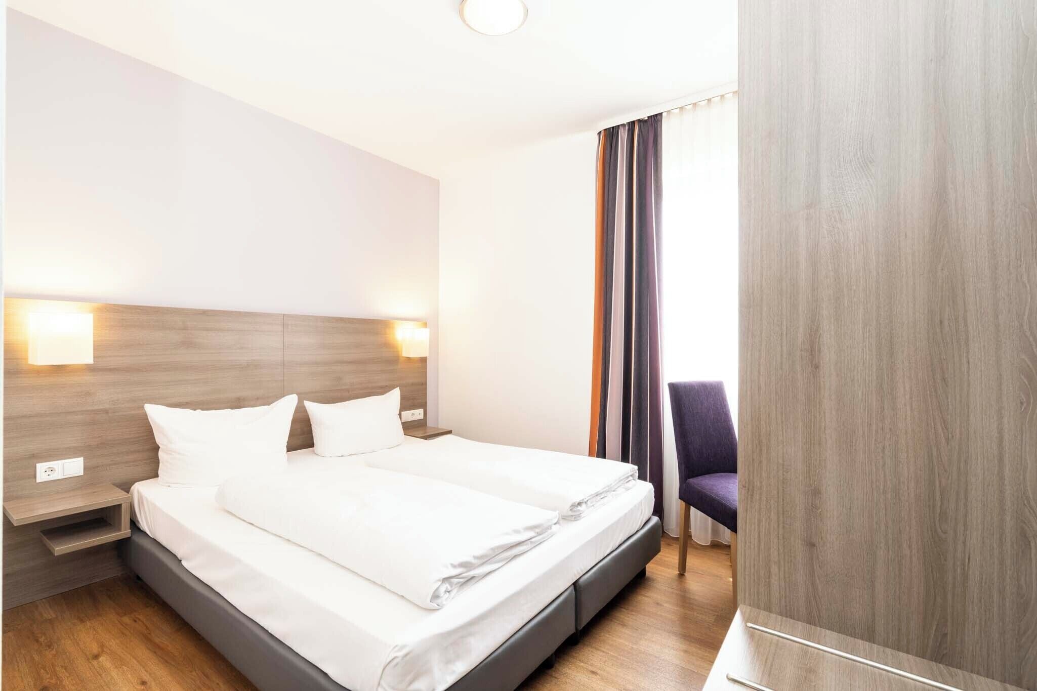 Ein Hotelzimmer mit Doppelbett, Holz-Kopfteil, Nachttischlampen, einem Sessel und Blick durch Vorhänge nach draußen.
