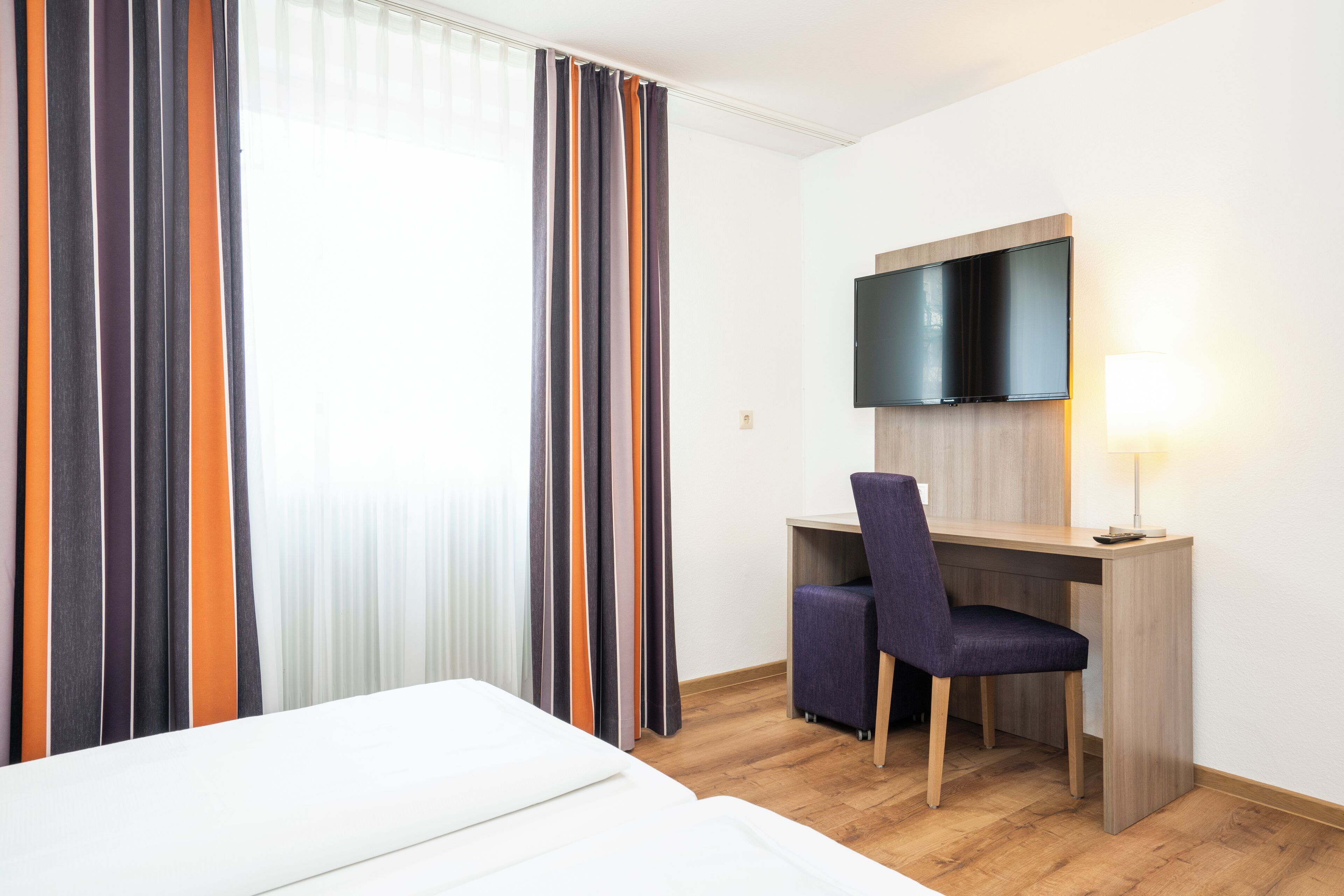 A hotel room with a bed, a desk with a chair, a TV mounted on the wall, and curtains.