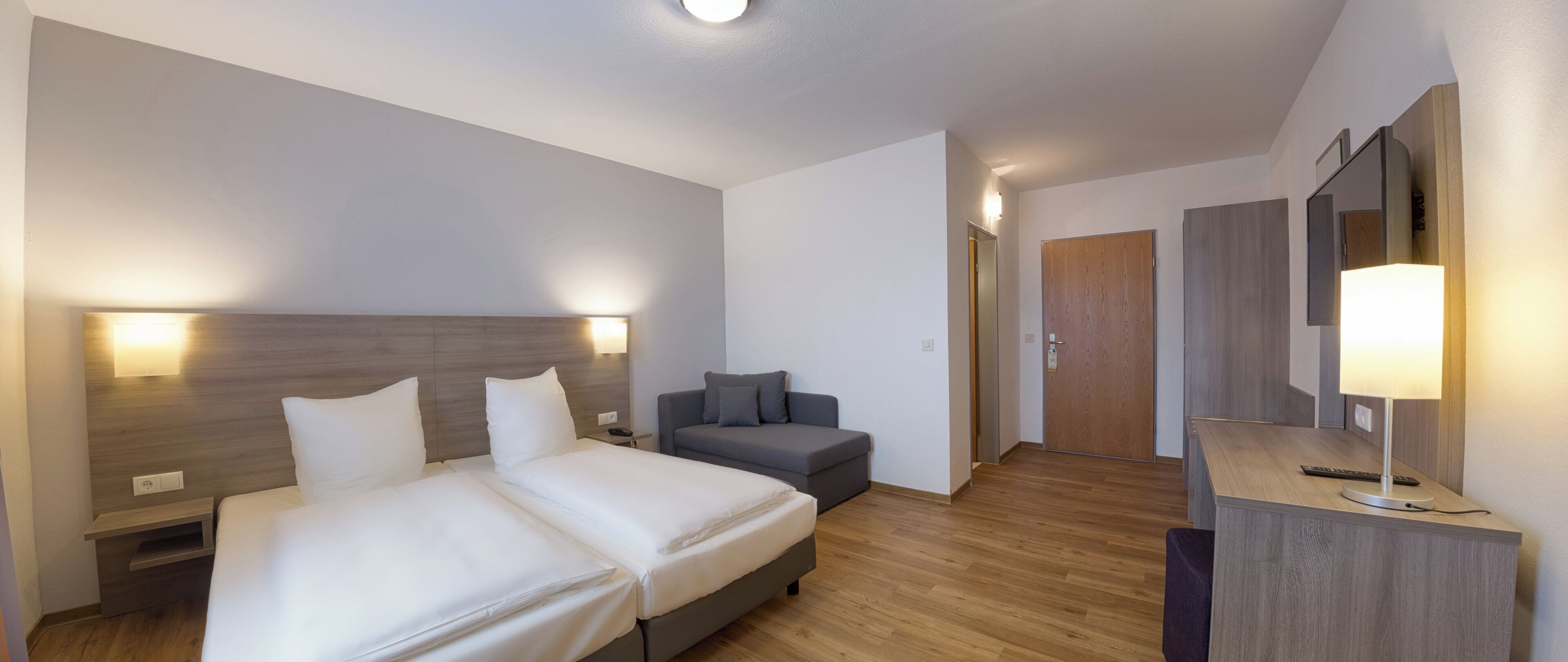 A hotel room with two beds, a sofa, a desk with a lamp, and a door leading to another room.