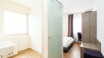 A modern hotel room with a glass shower enclosure, a bed with a wooden headboard, a small seating area, and a window providing natural light.