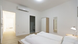 Un dormitorio moderno con dos camas, piso de madera, paredes blancas, un equipo de aire acondicionado y una lámpara de noche.