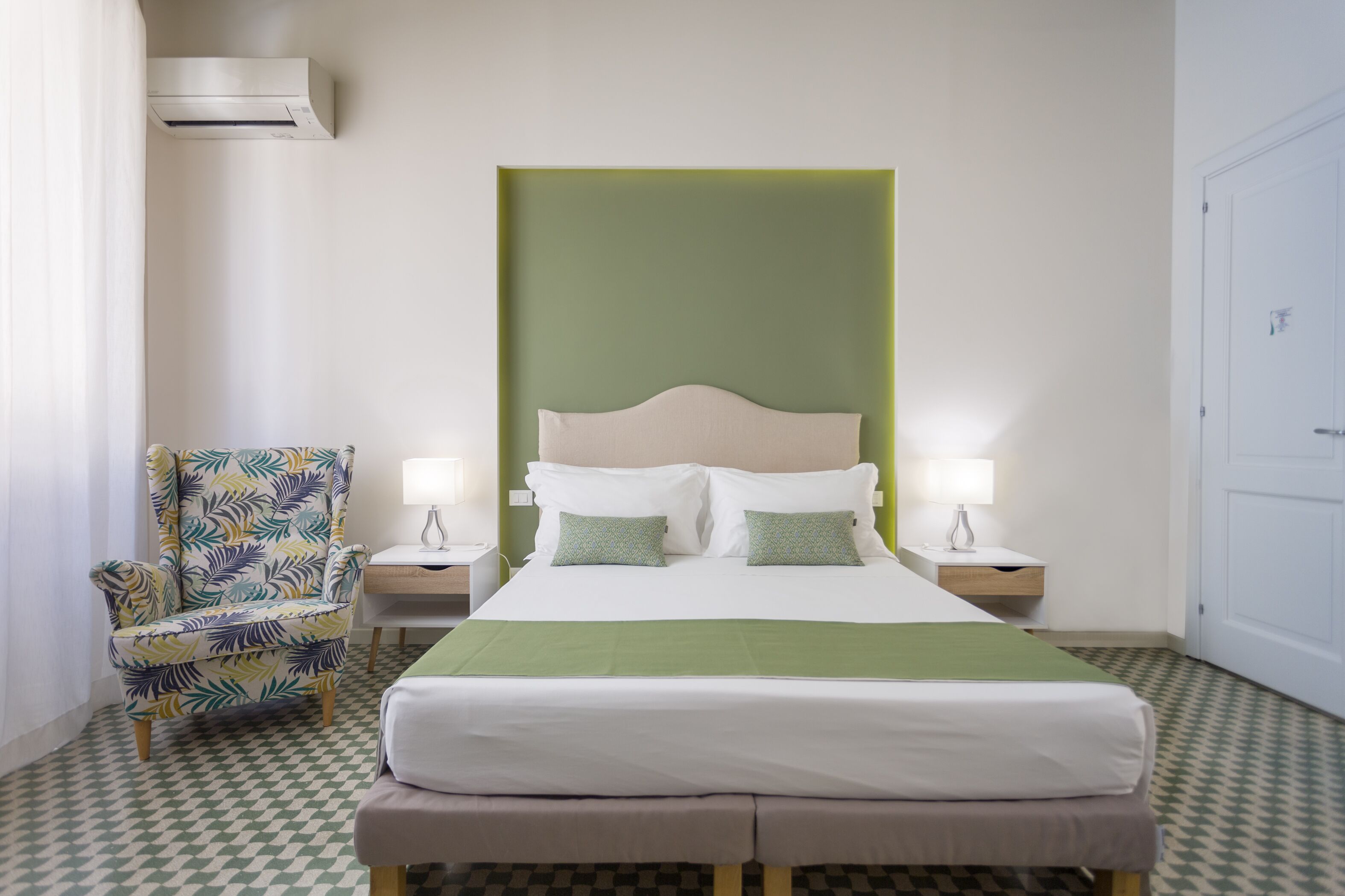 Habitación de hotel con una cama, una silla, dos mesitas de noche con lámparas y un equipo de aire acondicionado.
