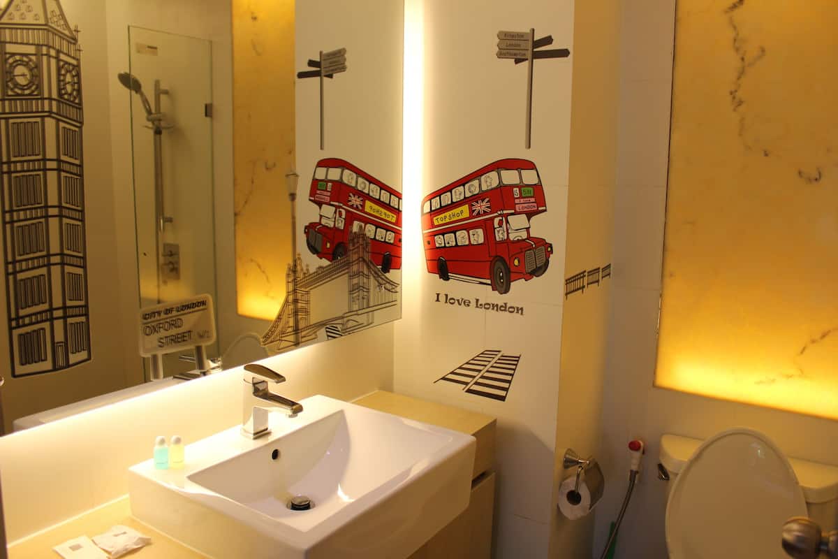 A bathroom with a sink, mirror, and toilet. There are wall decorations including a clock tower and a red double-decker bus.