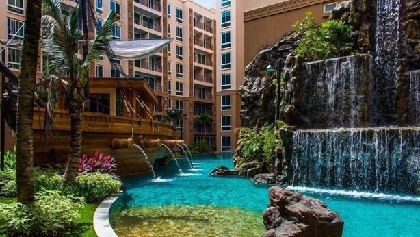 Una piscina de hotel con cascada, palmeras y un edificio de varios pisos al fondo.