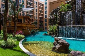 Una piscina de hotel con cascada, palmeras y un edificio de varios pisos al fondo.