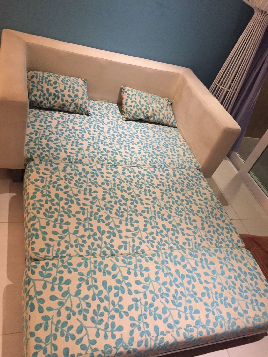 Een bed met een sprei met bladmotief in een kamer met een blauwe wand en een raam met een gordijn.