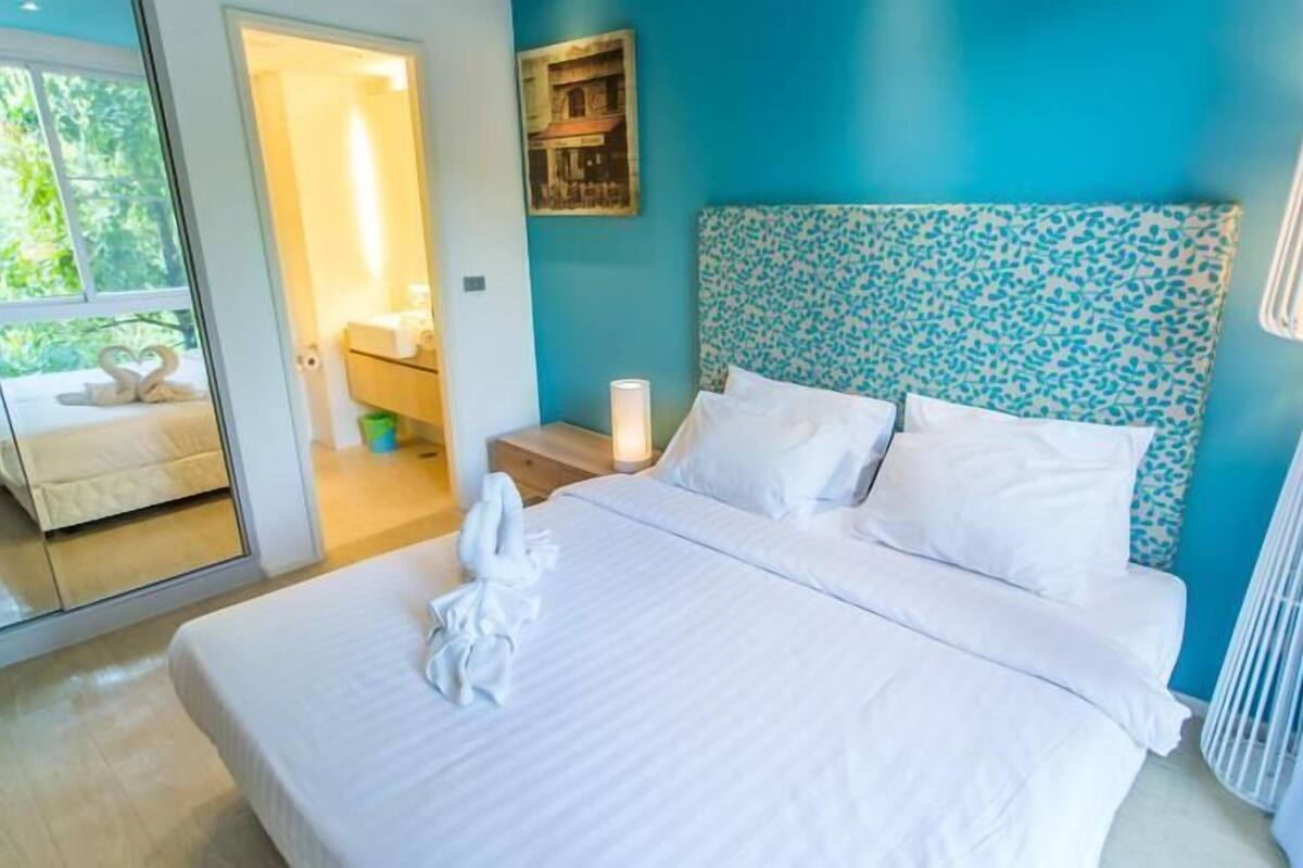 A bedroom with a bed, a nightstand, a lamp, a mirror, and a view of the outside.
