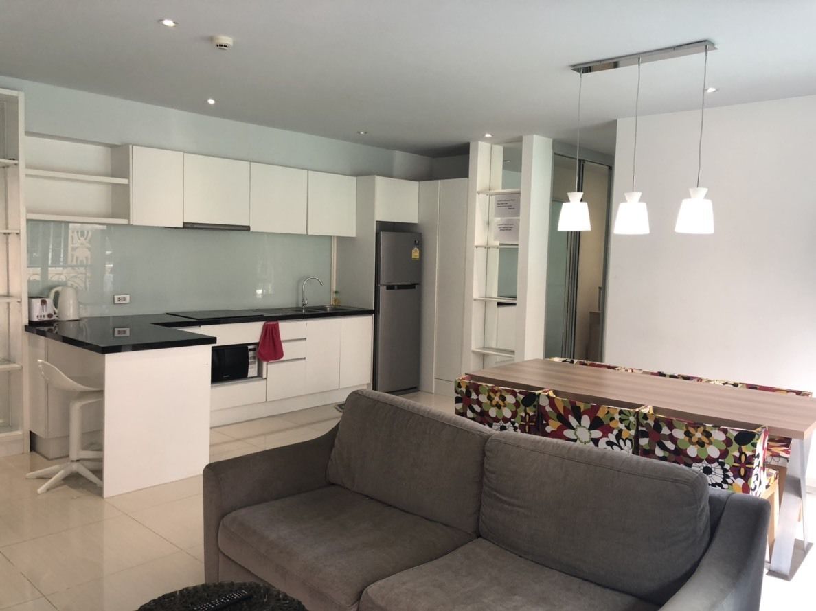 A modern kitchen with white cabinets, a black countertop, and a built-in oven. There is a gray sofa and a colorful table in the living area.