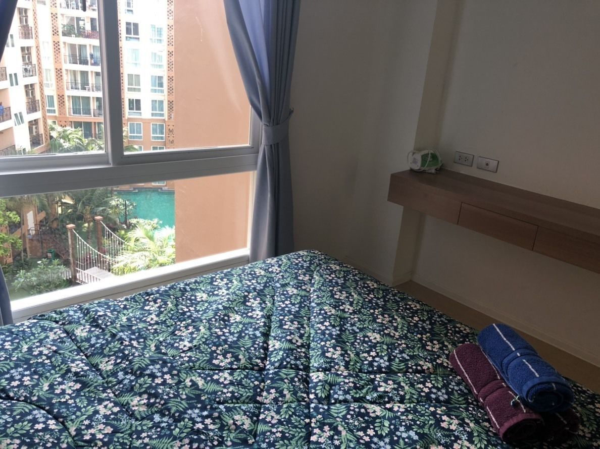 A hotel room with a bed, a window with curtains, a wooden headboard, and a small shelf with a clock and a power outlet.