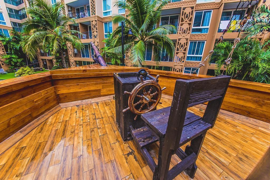 A wooden deck with a ship's wheel and a chair, surrounded by lush greenery and a multi-story building with balconies.