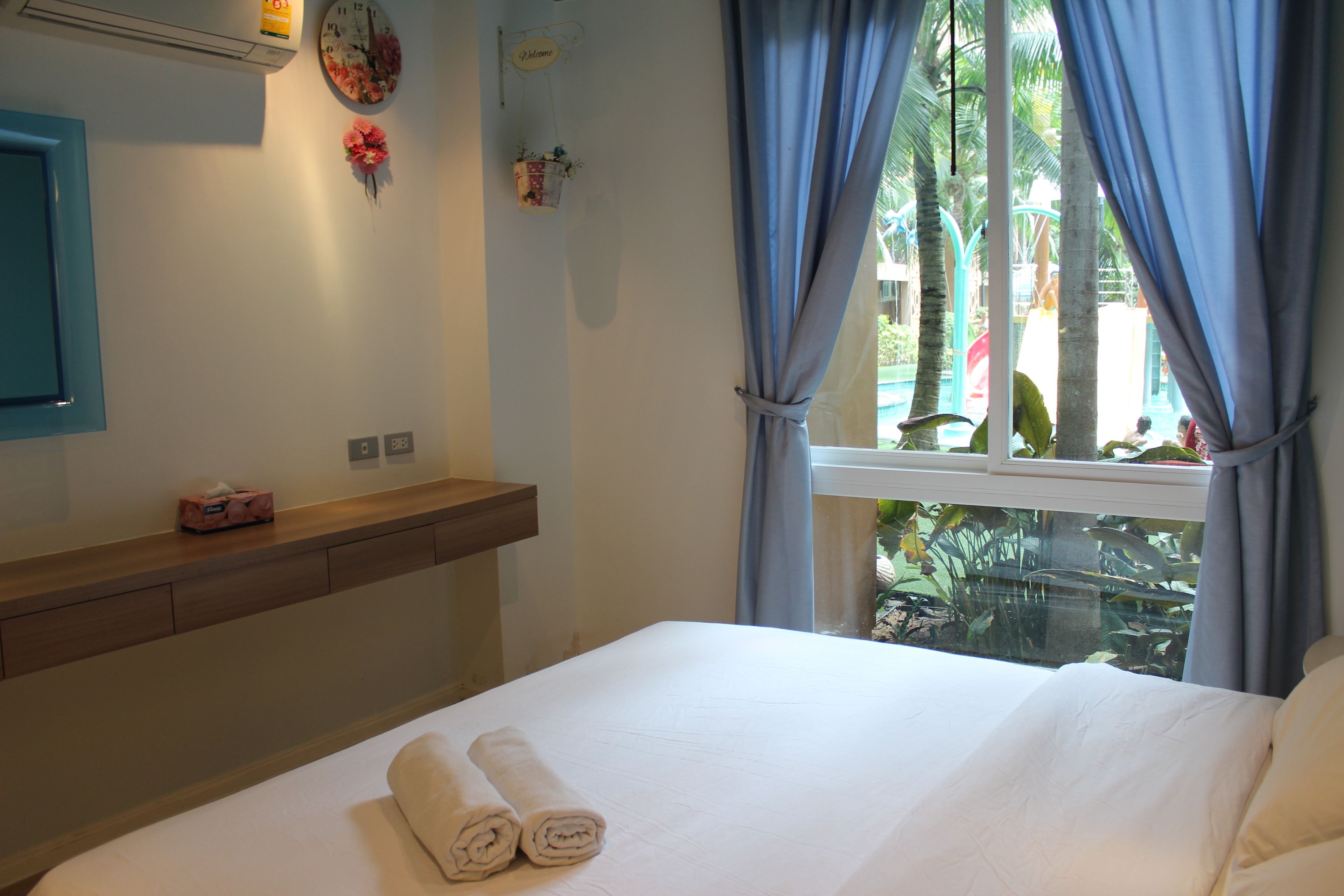 Quarto de hotel com cama, mesa de cabeceira, relógio e janela com cortinas.
