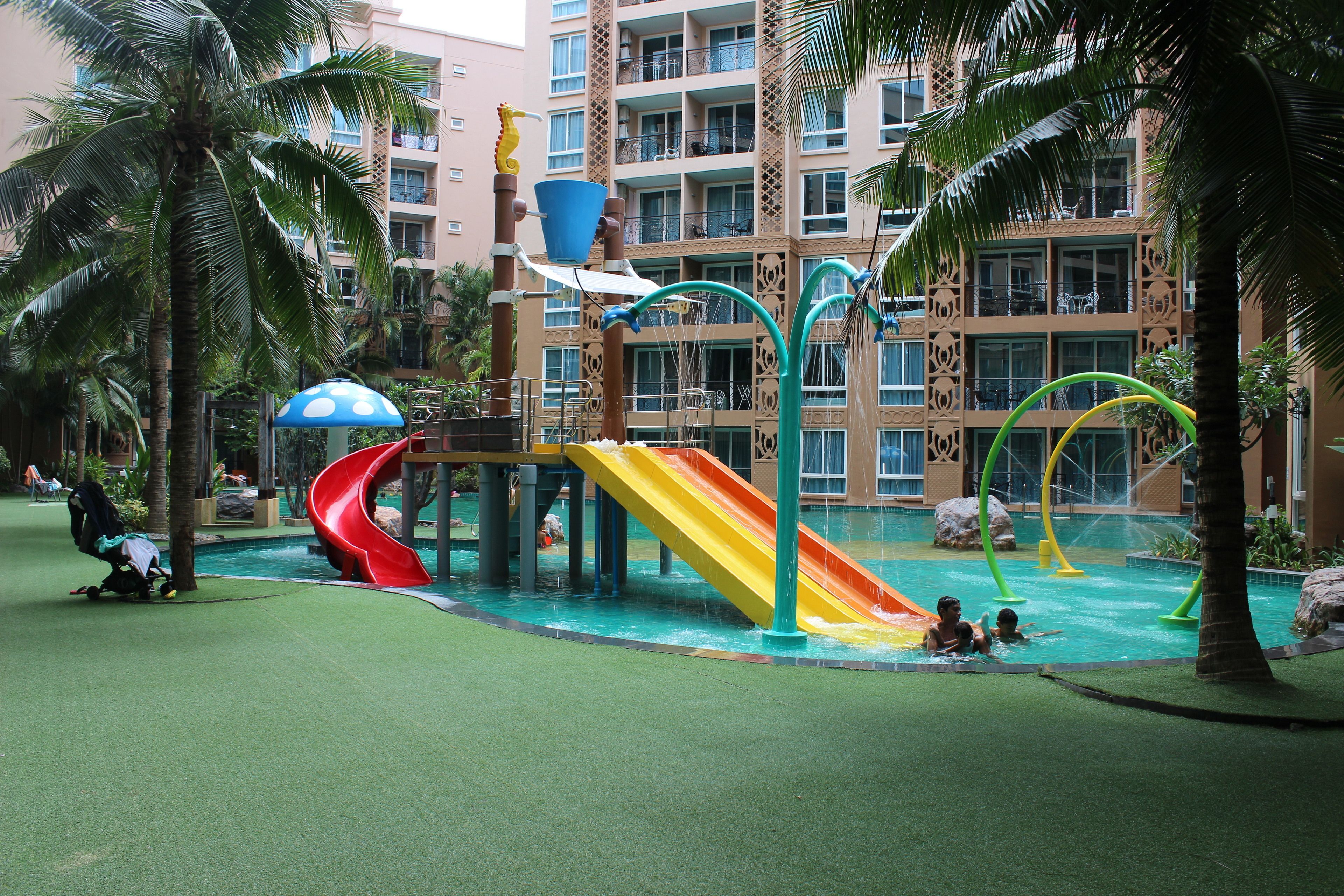 A water park with slides and pools, surrounded by palm trees and a hotel building in the background.