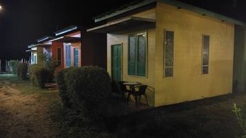 Un edificio pequeño de una sola planta con techo plano y un patio cubierto. Hay una mesa y sillas afuera, y se ve una puerta verde.