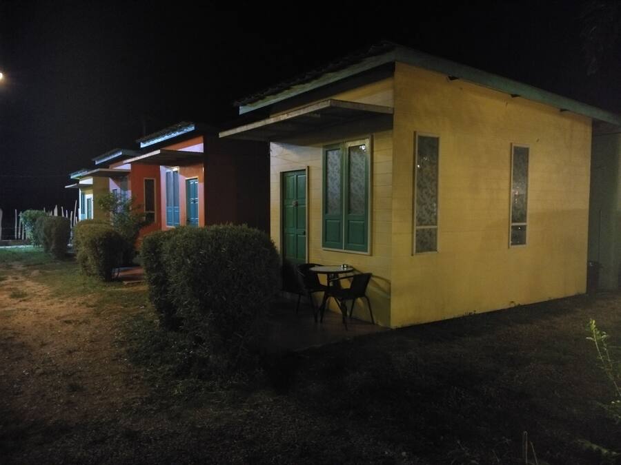 Un edificio pequeño de una sola planta con techo plano y un patio cubierto. Hay una mesa y sillas afuera, y se ve una puerta verde.