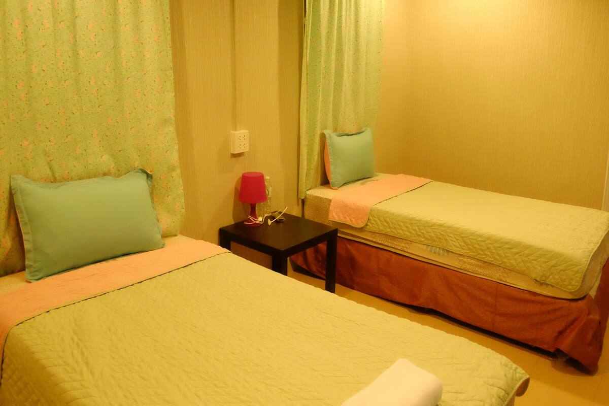 Una habitación con dos camas, cada una con una almohada y una manta. Hay una pequeña mesa con una lámpara y un objeto rojo encima.