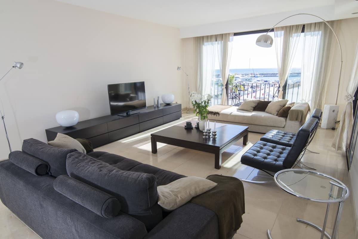 A modern living room with a dark grey sofa, a black leather chair, a glass coffee table, and a flat-screen TV on a black stand.