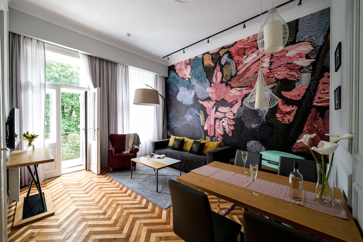 A modern living room with a herringbone floor, a sofa with colorful cushions, a dining table with chairs, and a large floral mural on the wall.
