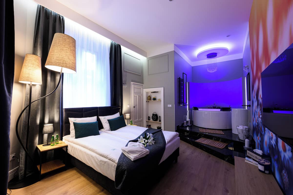 A modern hotel room with a large bed, bedside lamps, a small table with bottles, and a bathroom with a bathtub and shower.